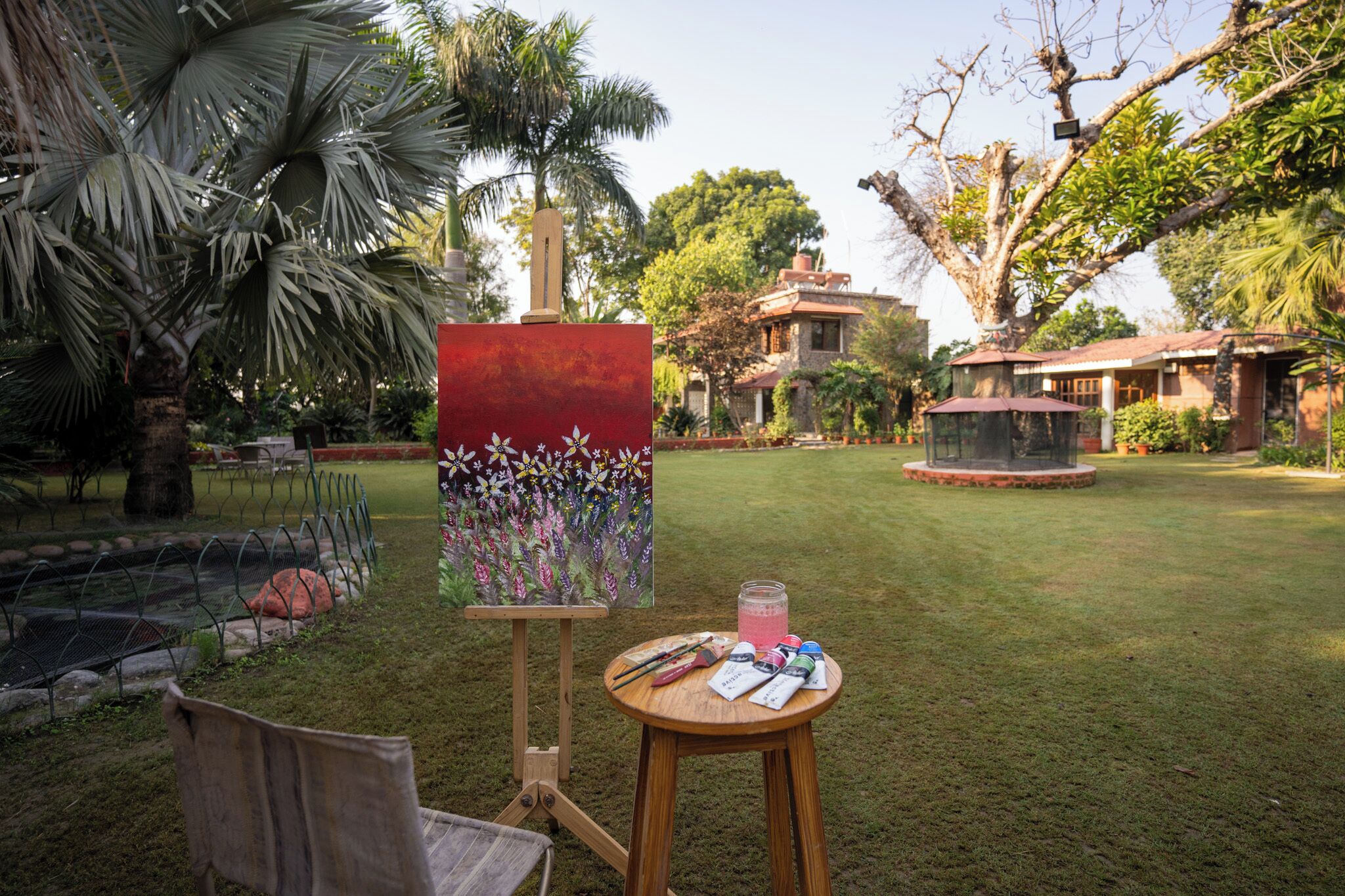 An outdoor painting setup with a canvas, easel, and art supplies on a table, surrounded by a well-maintained garden and a house in the background.