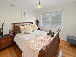 A bedroom with a wooden bed, a bedside table, a nightstand, a window with blinds, and a hanging light fixture.