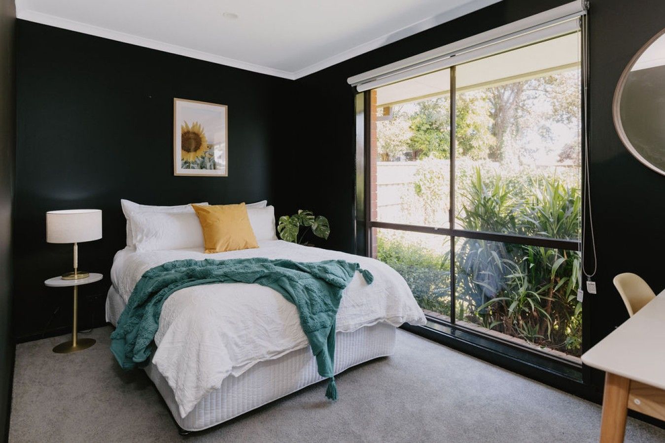 Quarto com cama, luminária de cabeceira, janela com vista para área verde e um quadro na parede.