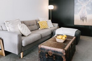 A living room with a grey sofa, patterned cushions, a lamp, a painting of a deer, and a vintage suitcase with fruit on top. - 2 King Beds | Walk to Town | Sleeps 6 (Sale)