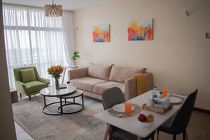 Un saló modern amb un butac armchair verd, un sofà beix i una taula de dinar preparada per a dues persones. Hi ha dos quadres abstractes a la paret, un vas amb flors damunt la taula de centre i una planta en test.