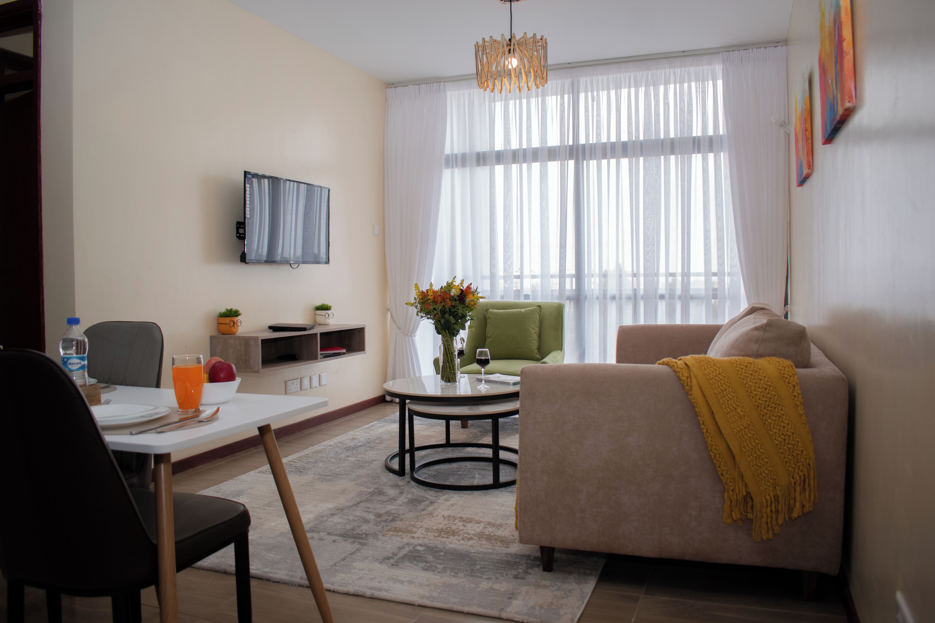 Una sala moderna con un sofá, una mesa de comedor con sillas, un televisor montado en la pared y un ventanal con cortinas translúcidas.