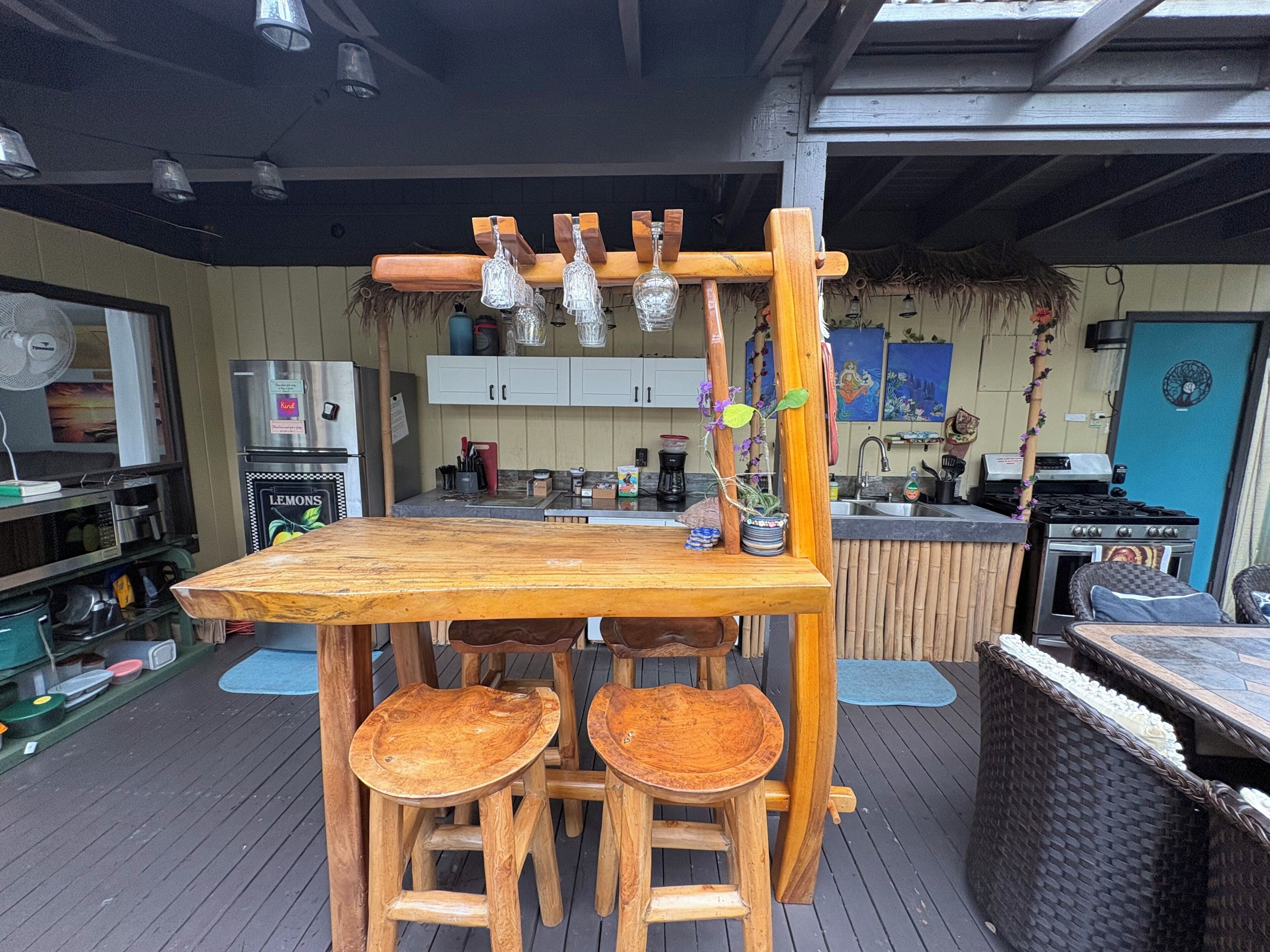 Una cocina exterior de madera con barra y taburetes, una lámpara colgante y una puerta azul al fondo.