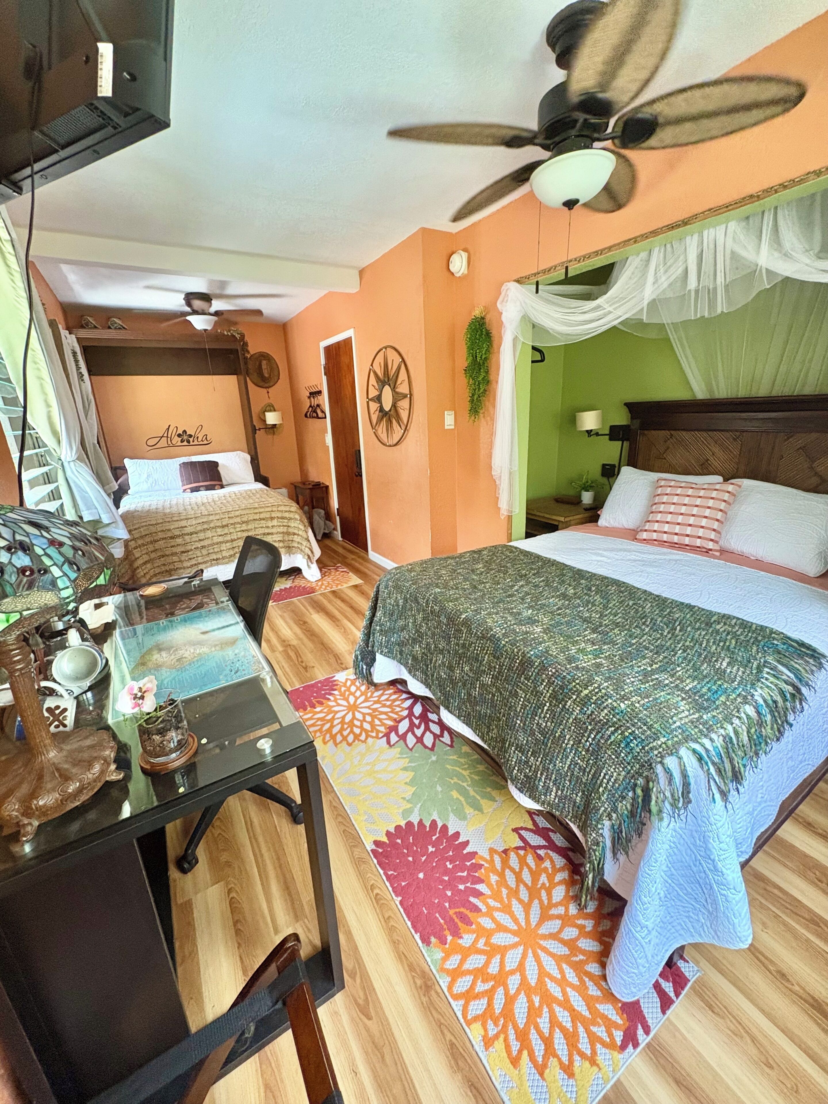 A bedroom with a bed, a desk, a chair, a ceiling fan, and a nightstand with a lamp.