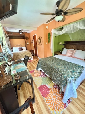 A bedroom with a bed, a desk, a chair, a ceiling fan, and a nightstand with a lamp.