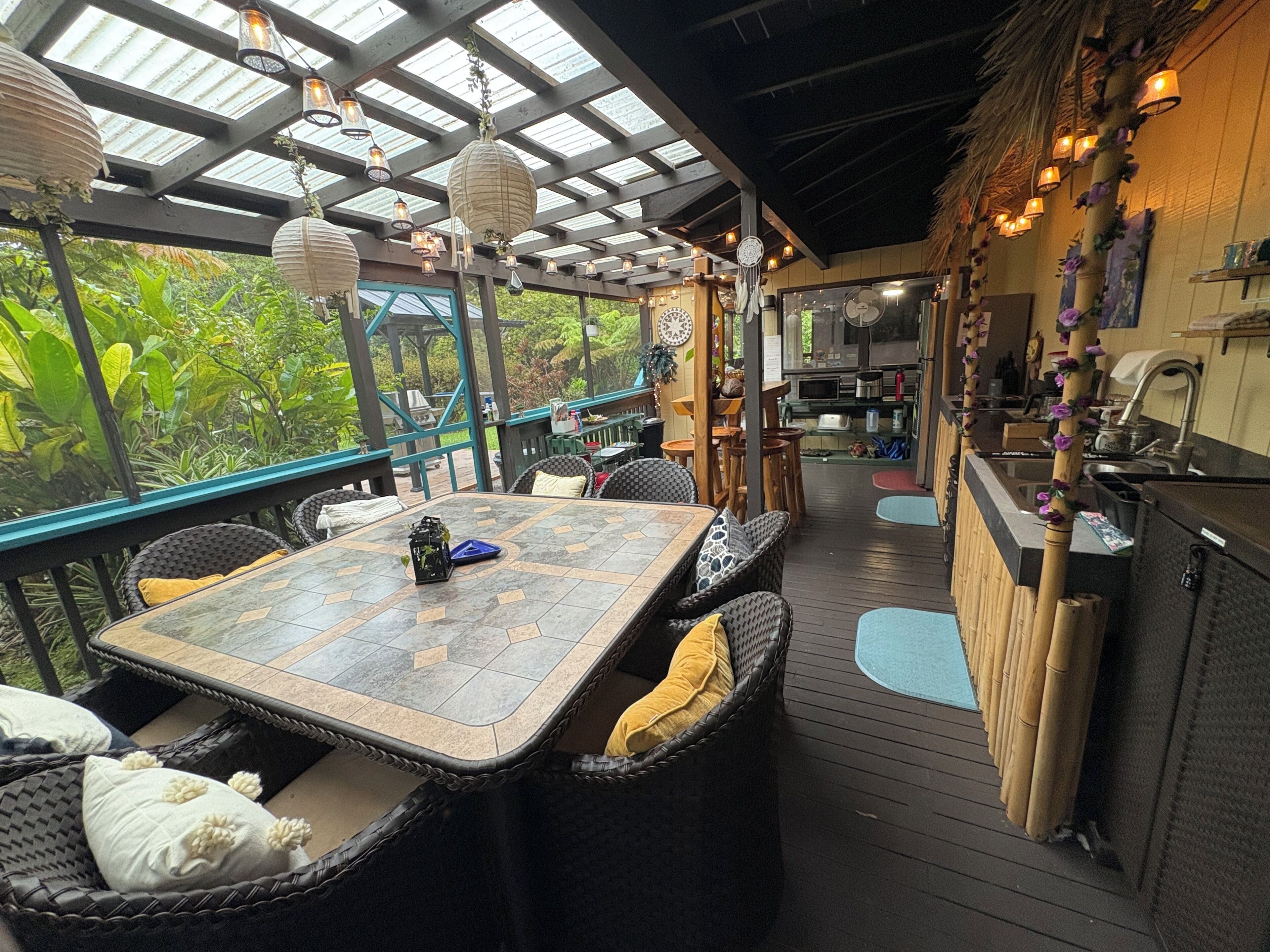 An outdoor dining area with a table and chairs, a kitchen area, and a view of greenery.
