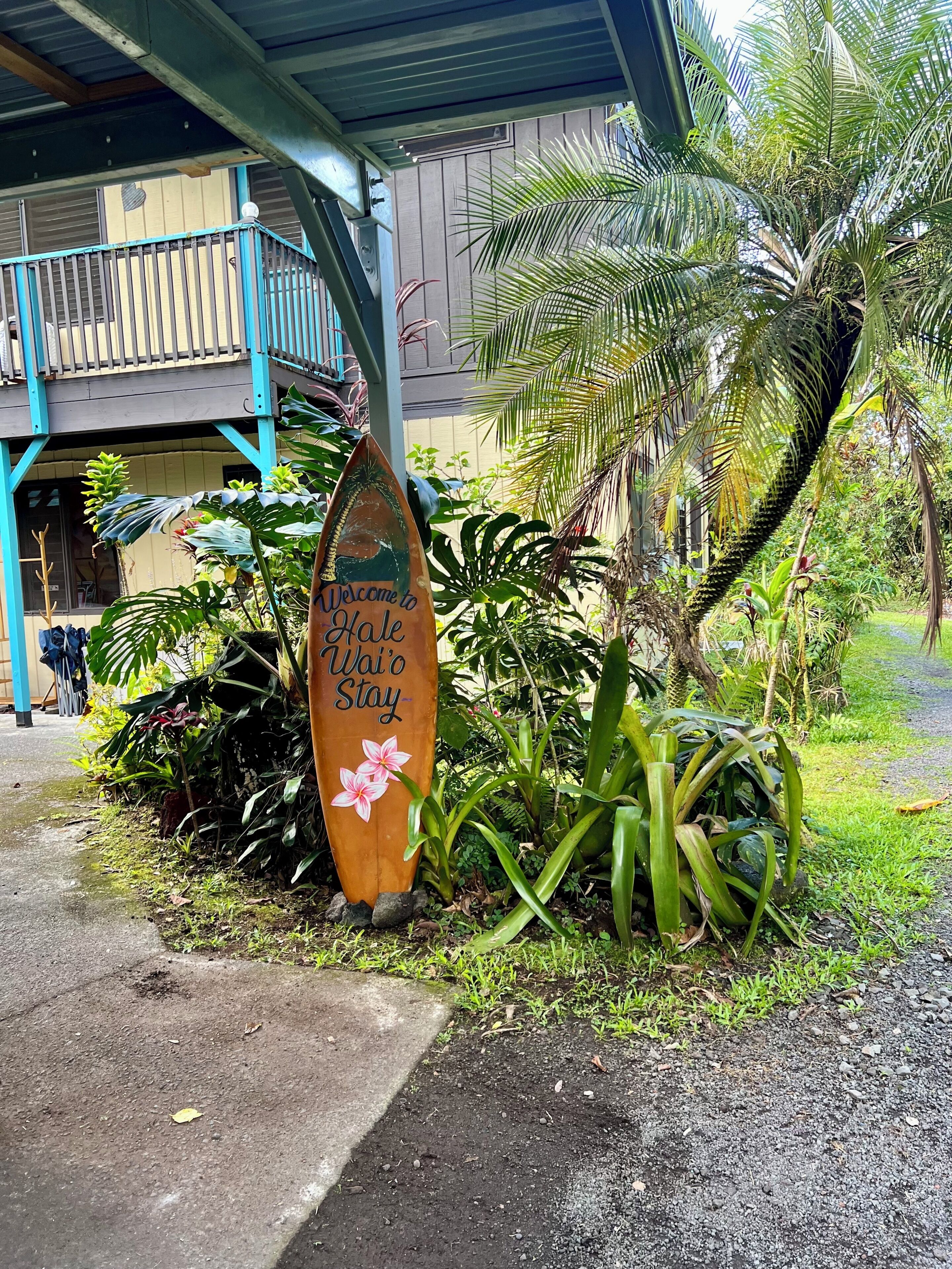 A wooden sign with 'Welcome to Wai'o Stay' written on it, placed in front of a building with a blue balcony and tropical plants.