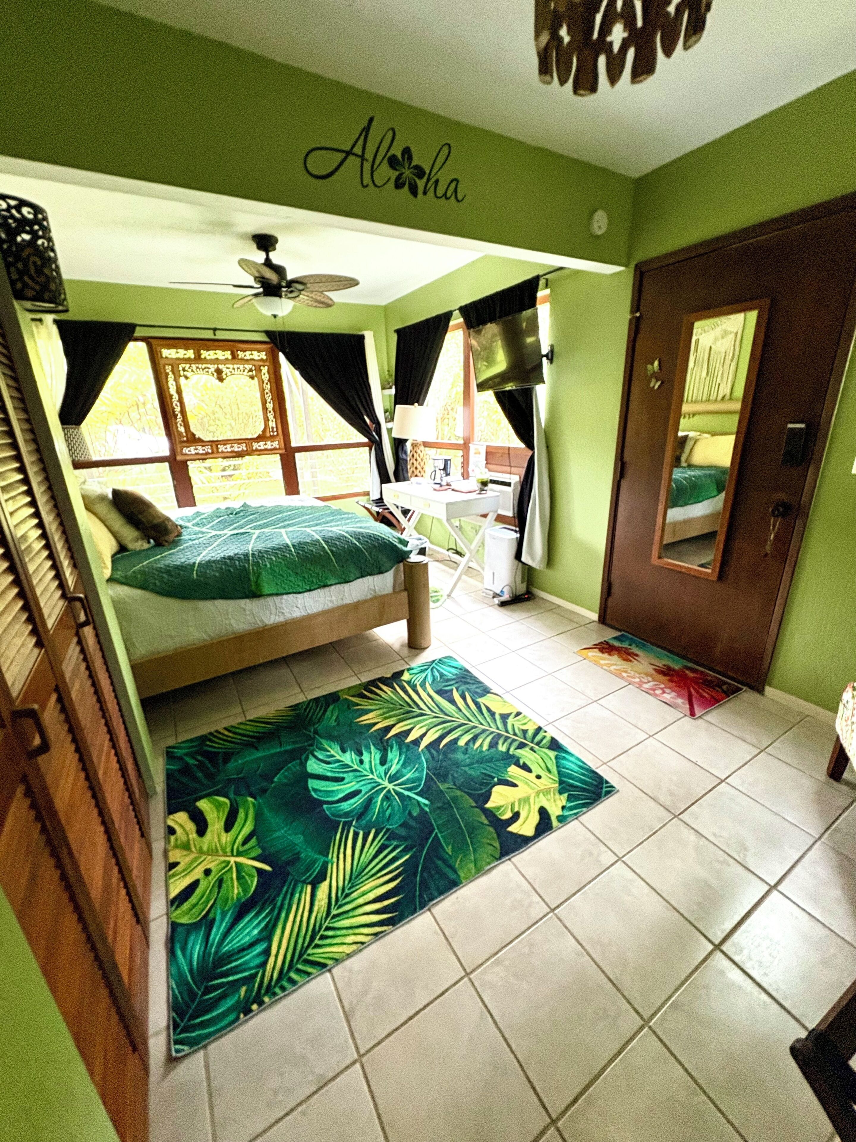 Une chambre comprenant un lit, un bureau, une chaise, un miroir, un ventilateur de plafond et un tapis à motifs tropicaux.
