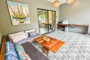 A living room with a sofa, a wooden coffee table, a painting of boats, a sliding glass door leading to a balcony, and a kitchen area with a microwave and sink.