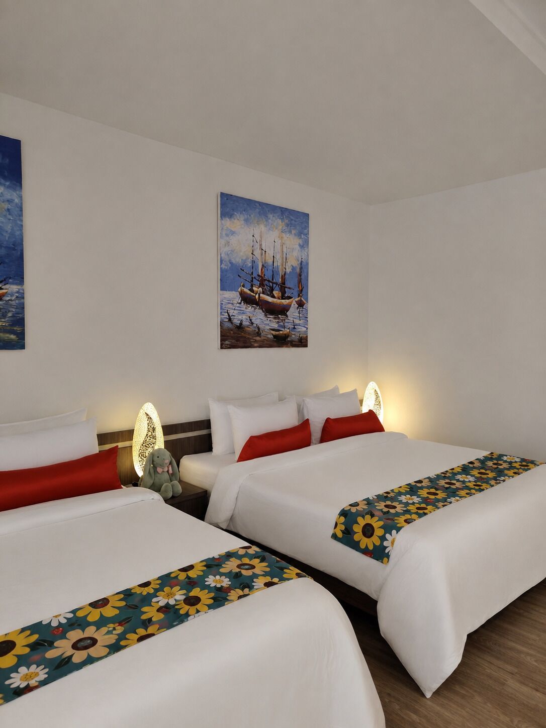 A hotel room with two beds, white bedding, red pillows, a floral bedspread, a painting of boats on the wall, and a nightstand with a lamp.