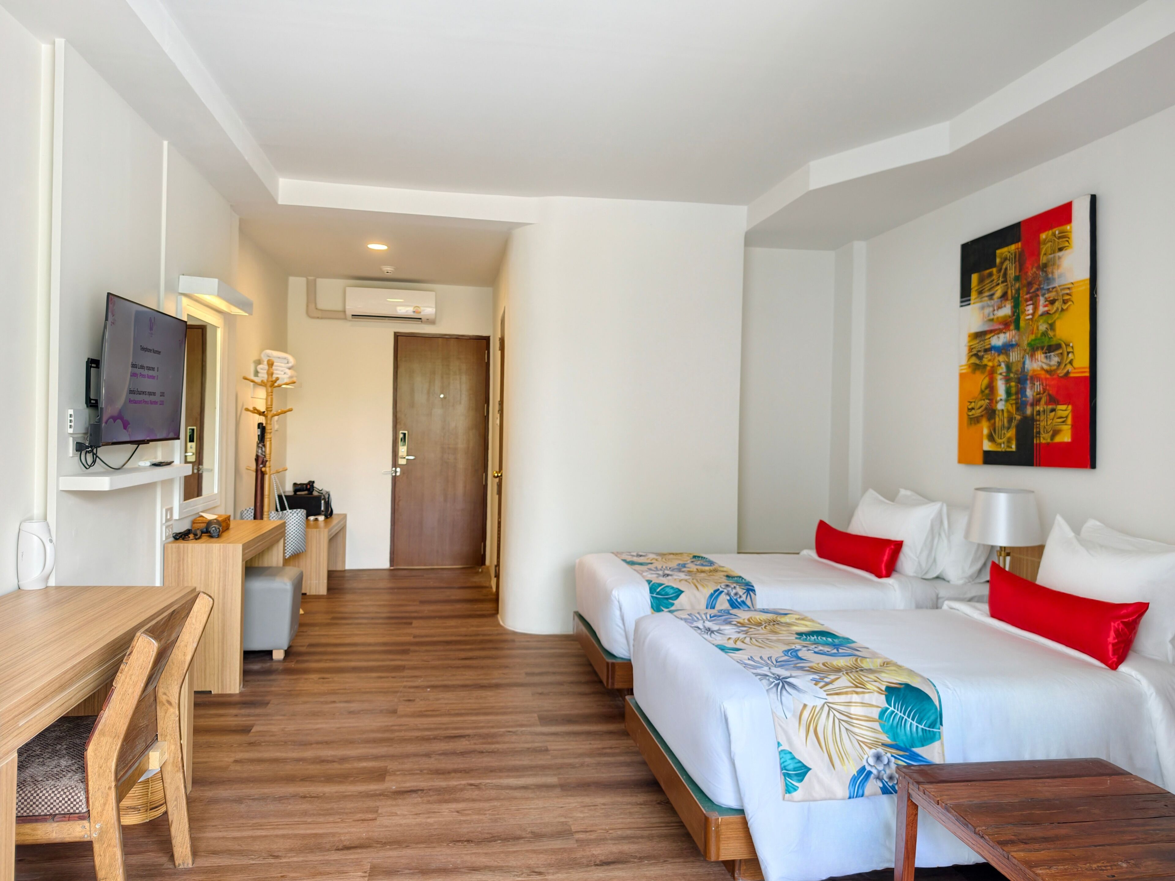 Quarto de hotel com escrivaninha de madeira, duas camas com colchas florais, uma televisão e um quadro na parede.