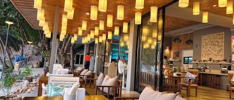 An indoor dining area with wooden ceiling and hanging lamps, a seating arrangement with tables and chairs, and a view of the outdoor area through large windows.