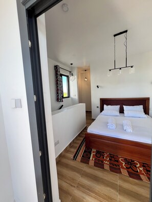 A hotel room with a bed, a bedside table, a window with curtains, a wall-mounted light fixture, and a patterned rug. - Kameleon Villa’s (Kiwengwa)