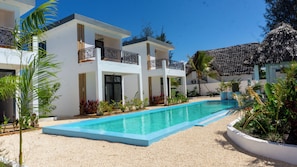 A modern two-story building with a swimming pool, surrounded by tropical plants and a thatched-roof structure. - Kameleon Villa’s (Kiwengwa)