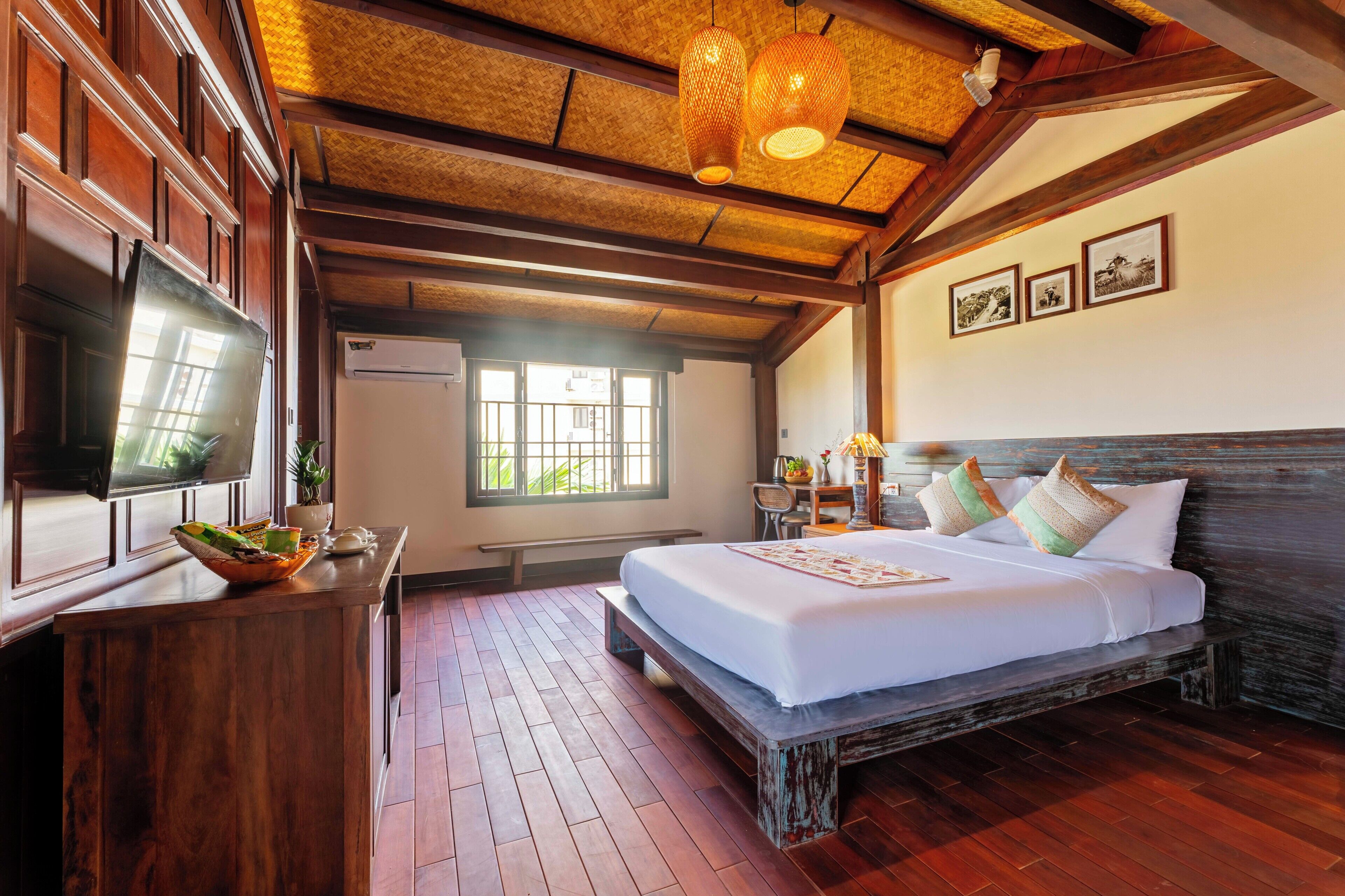 A hotel room with a wooden bed, a television, a desk with a plant, and a window with a view of greenery.