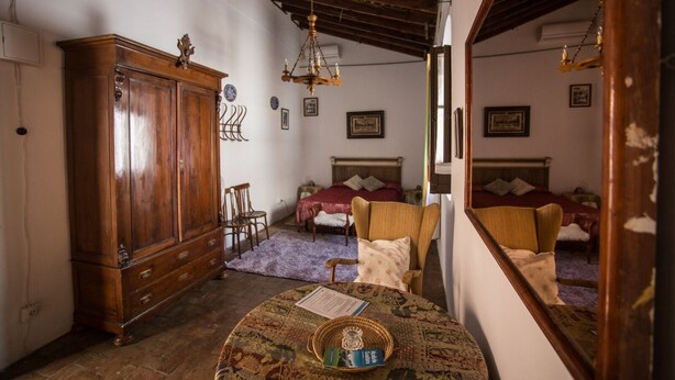 A room with a wooden wardrobe, a bed with a red bedspread, a chair, a small table with a basket and a book, and a mirror reflecting the room.