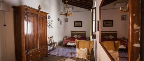 Una habitación con un armario de madera, una cama con colcha roja, una silla, una mesita con una cesta y un libro, y un espejo que refleja la habitación.