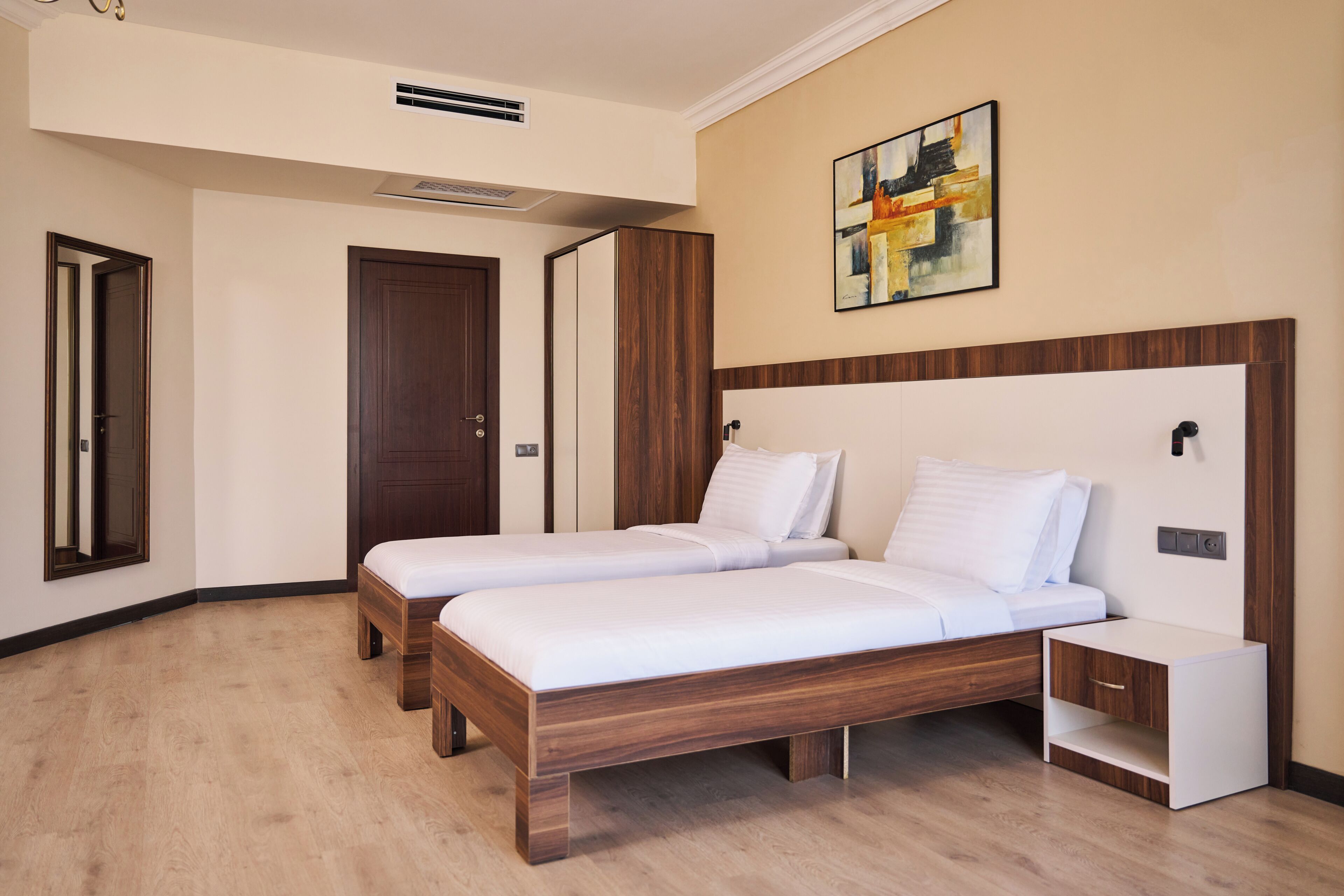 A hotel room with two beds, a wooden headboard, a bedside table, a nightstand, a painting on the wall, and a door.