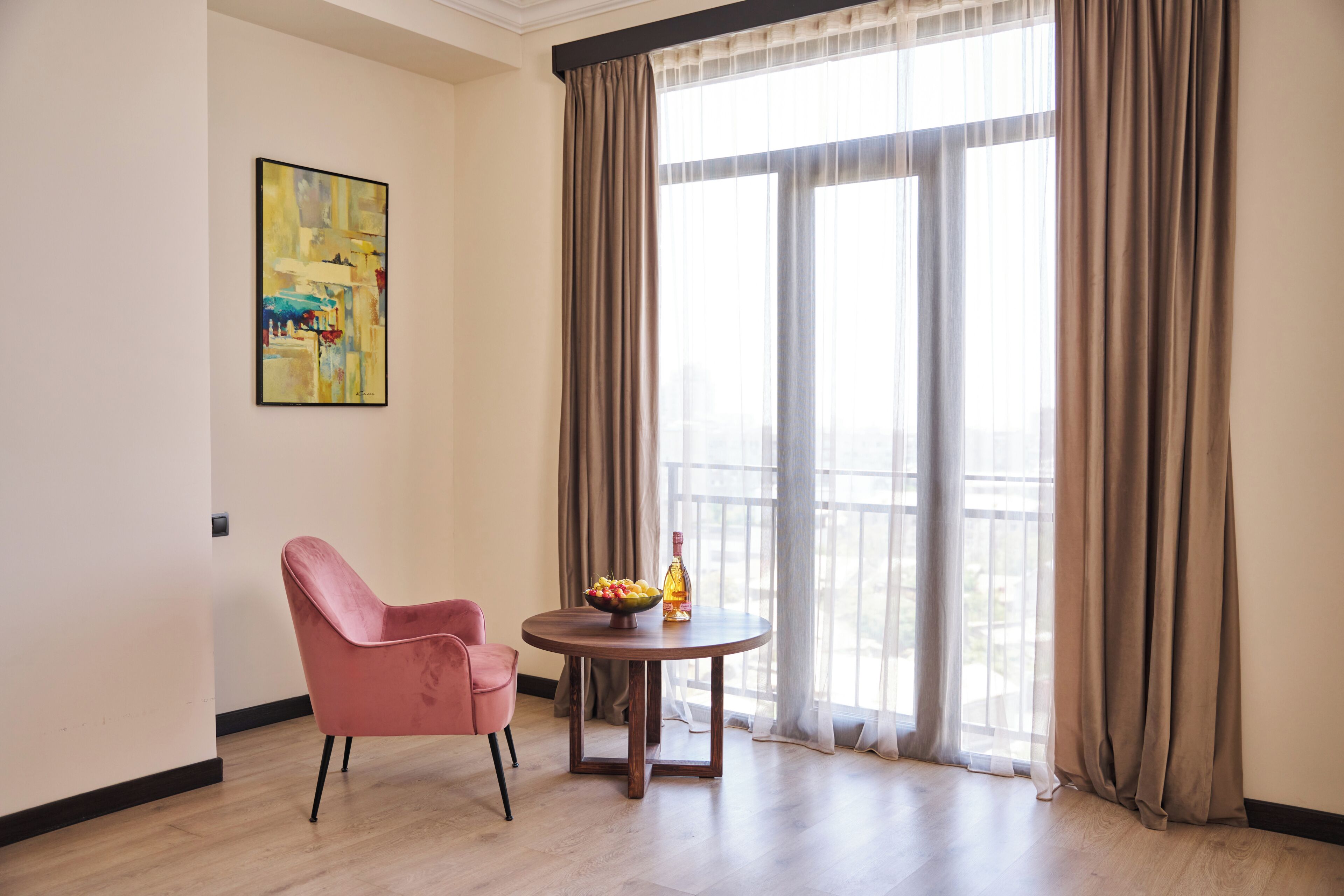 A room with a pink chair, a round wooden table with a bowl of fruit and a bottle, and a large window with curtains.