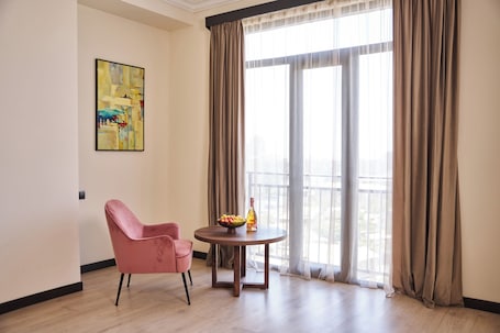 A room with a pink chair, a round wooden table with a bowl of fruit and a bottle, and a large window with curtains.