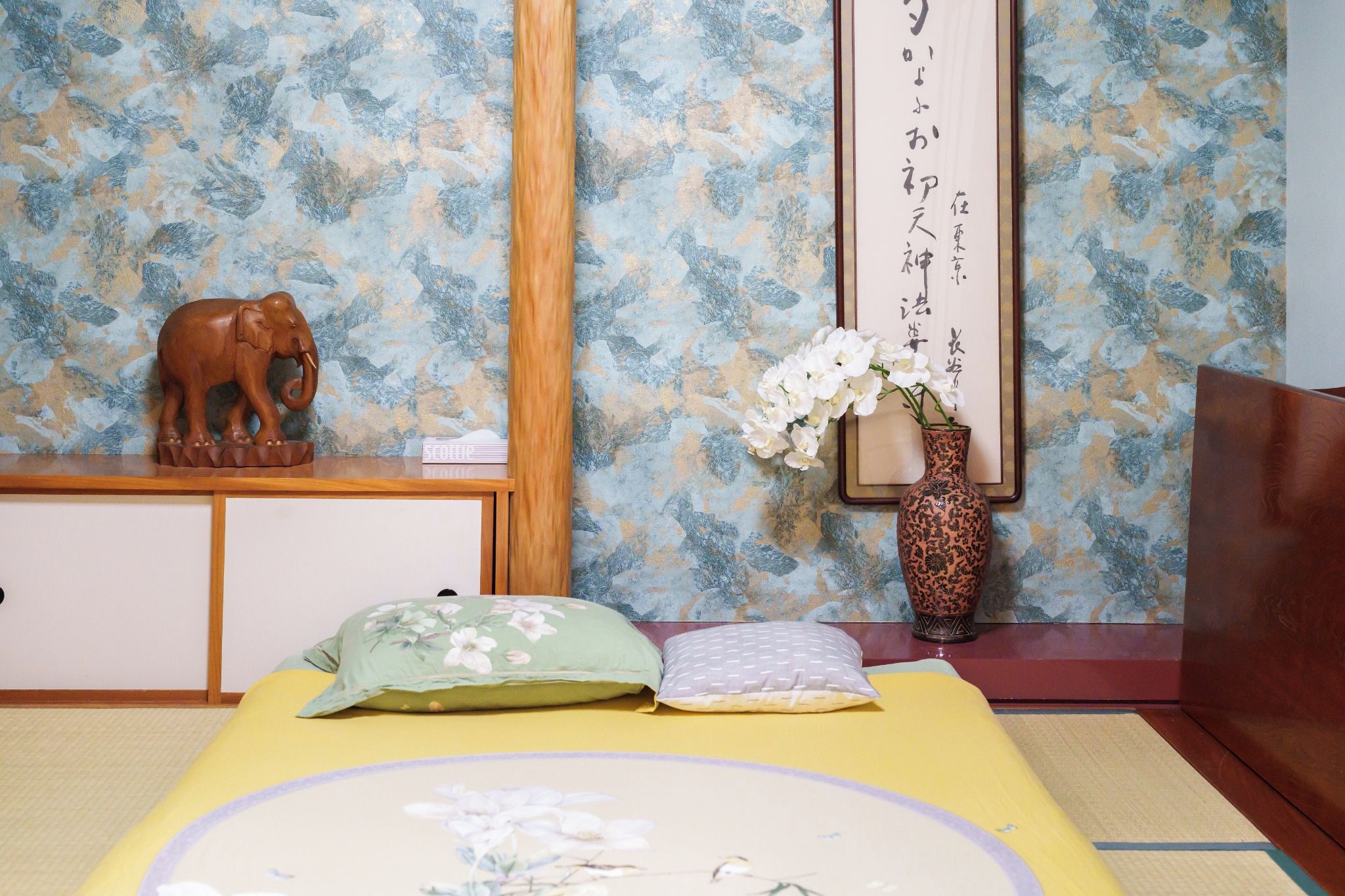 A traditional Japanese-style room with tatami flooring, a low wooden bed with a yellow bedspread, a wooden elephant sculpture, a vase with white flowers, and a framed calligraphy piece on the wall.