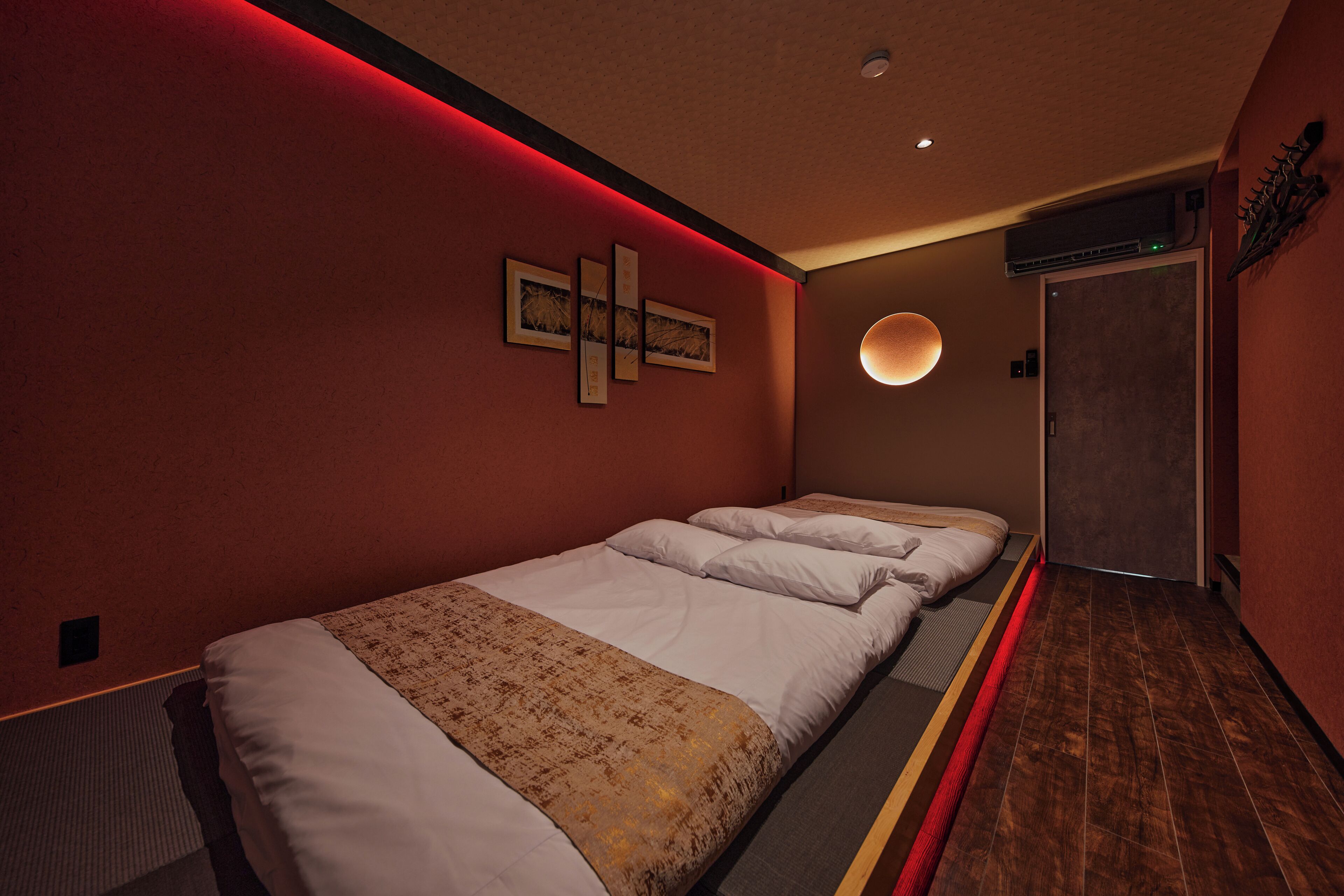 A hotel room with a long bed, a wall-mounted artwork, a round light fixture, and a door with a towel rack.