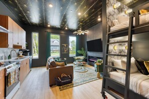 A modern living room with a sofa, coffee table, and a TV. There is a kitchen area with cabinets and a stove. The room has a unique ceiling design and large windows. - East Nashville 4-Home Buyout With Rooftop, Sleeps 33 (Nashville)