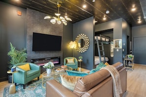 A modern living room with a flat-screen TV, a fireplace, a green armchair, a brown sofa, and a decorative mirror. - East Nashville 4-Home Buyout With Rooftop, Sleeps 33 (Nashville)