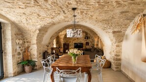 A stone-walled dining area with a wooden table and white chairs, a TV, and a fireplace in the background. - Stone house with a pool just outside Uzès (Garrigues-Sainte-Eulalie)