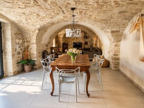 A stone-walled dining area with a wooden table and white chairs, a TV, and a fireplace in the background. - Stone house with a pool just outside Uz ès (Garrigues-Sainte-Eulalie)