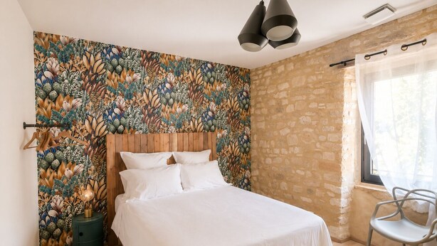 A bedroom with a bed, a headboard, a wall with a floral pattern, a lamp, a chair, and a window with curtains. - Stone house with a pool just outside Uzès (Garrigues-Sainte-Eulalie)
