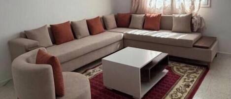 Una sala de estar con un sofá seccional color beige, una mesa de centro blanca y un tapete rojo estampado.