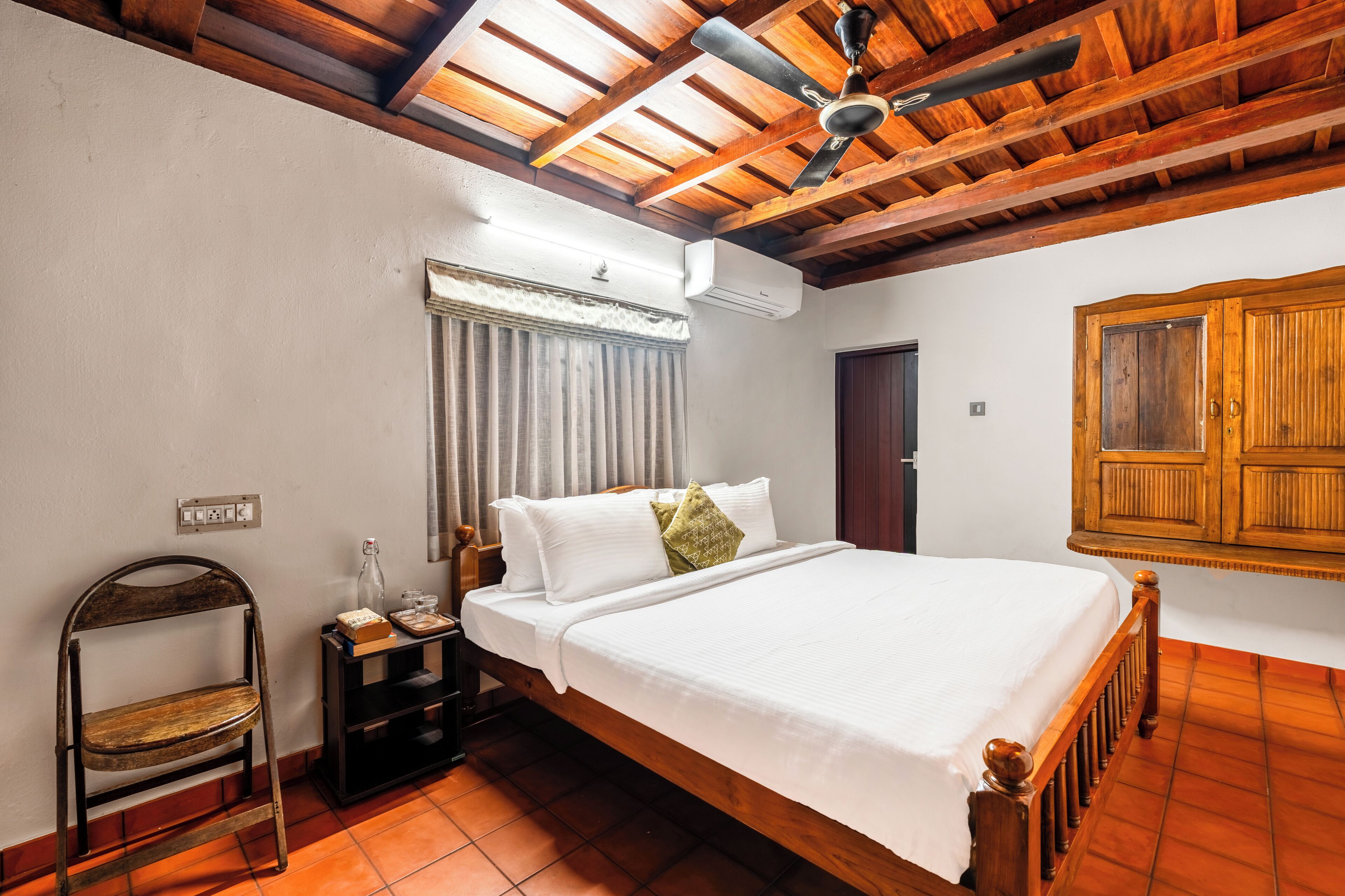 Un dormitorio con techo de madera, una cama con ropa blanca, una mesita de noche con una botella y vasos, una silla y una ventana con cortinas.