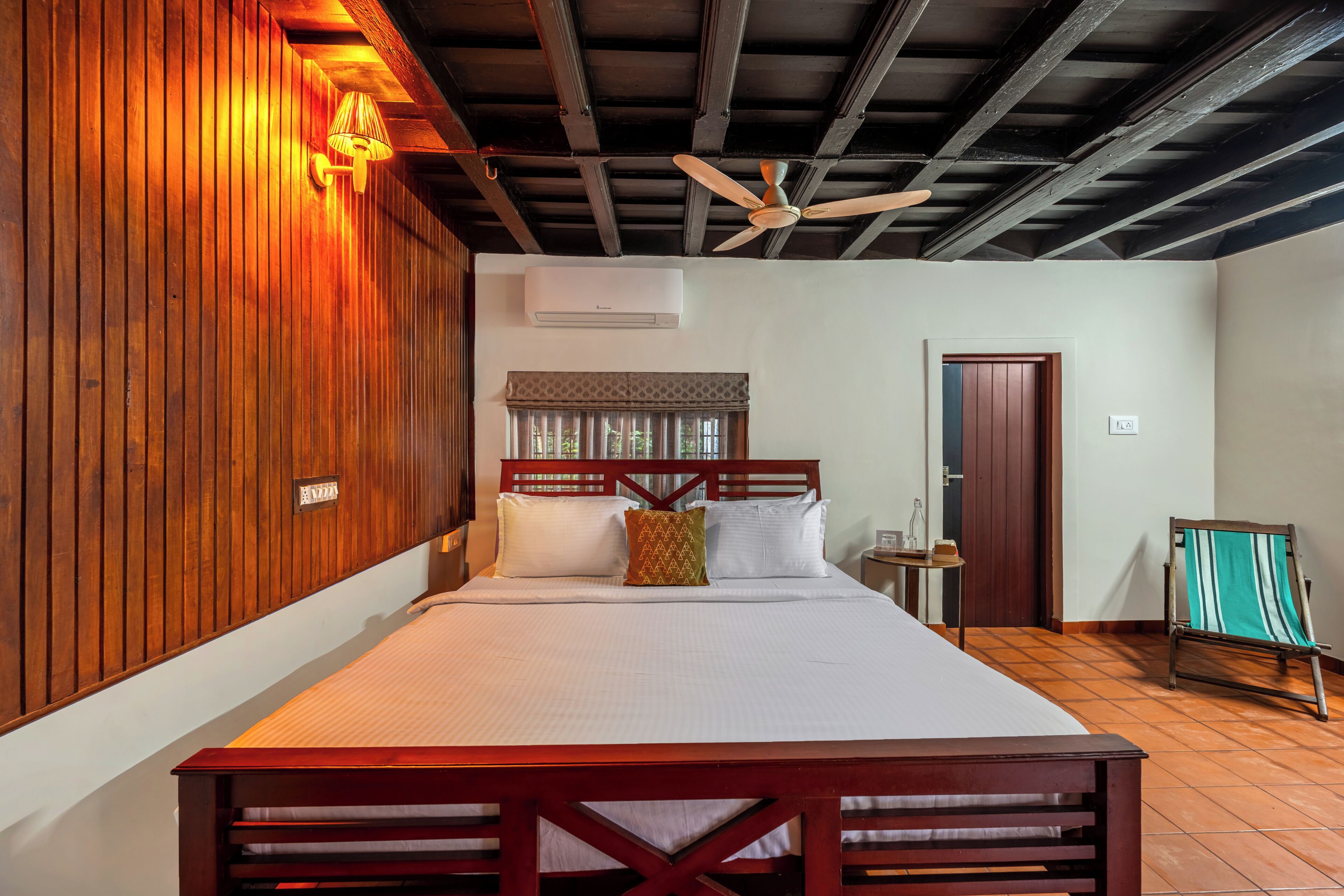 Habitación de hotel con cabecera de madera, cama con sábanas blancas, ventilador de techo y ventana con cortinas.