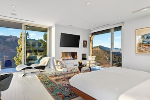 A modern bedroom with a large bed, a fireplace, a television, and a view of mountains through large windows.