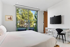 Een moderne hotelkamer met een groot bed, een bureau, een stoel, een televisie en een schuifdeur die leidt naar een balkon met uitzicht op bomen.