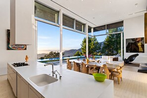A modern kitchen with a large island, a dining area with wooden chairs, and a view of mountains through large windows.