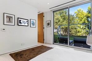 A modern bedroom with a large sliding glass door leading to a balcony with outdoor furniture.