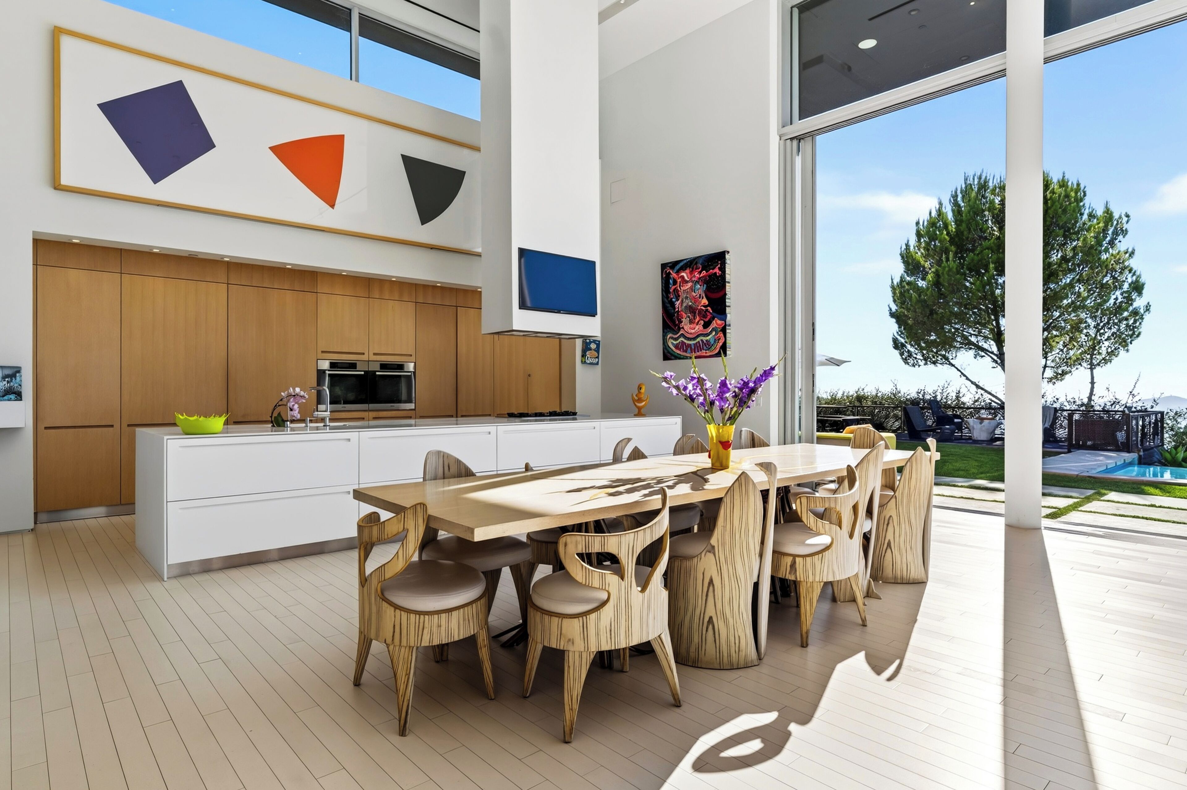 A modern kitchen with wooden cabinets, a dining area with a wooden table and chairs, and a large window overlooking a pool.