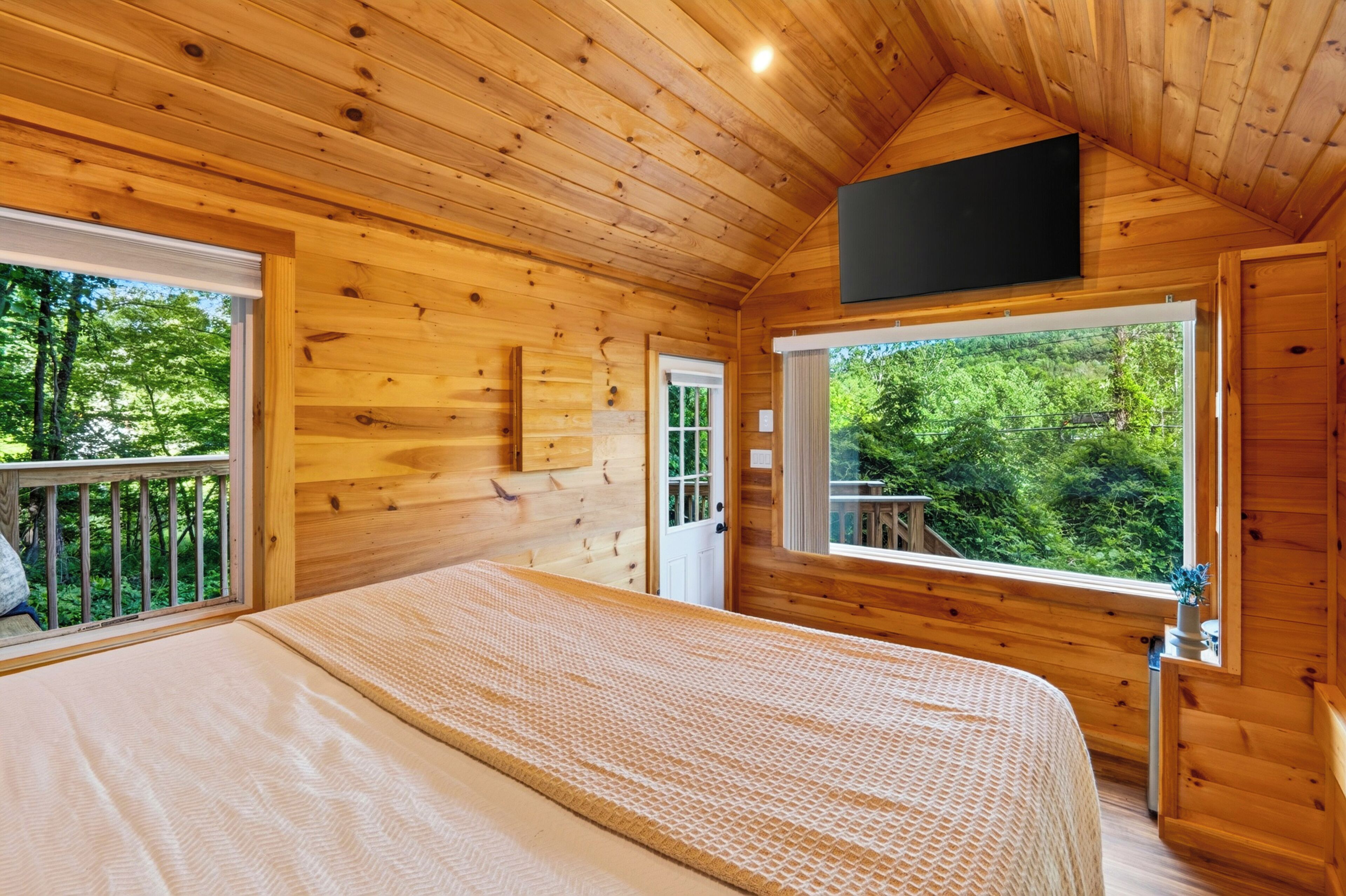 A wooden cabin bedroom with a bed, two windows, and a TV.