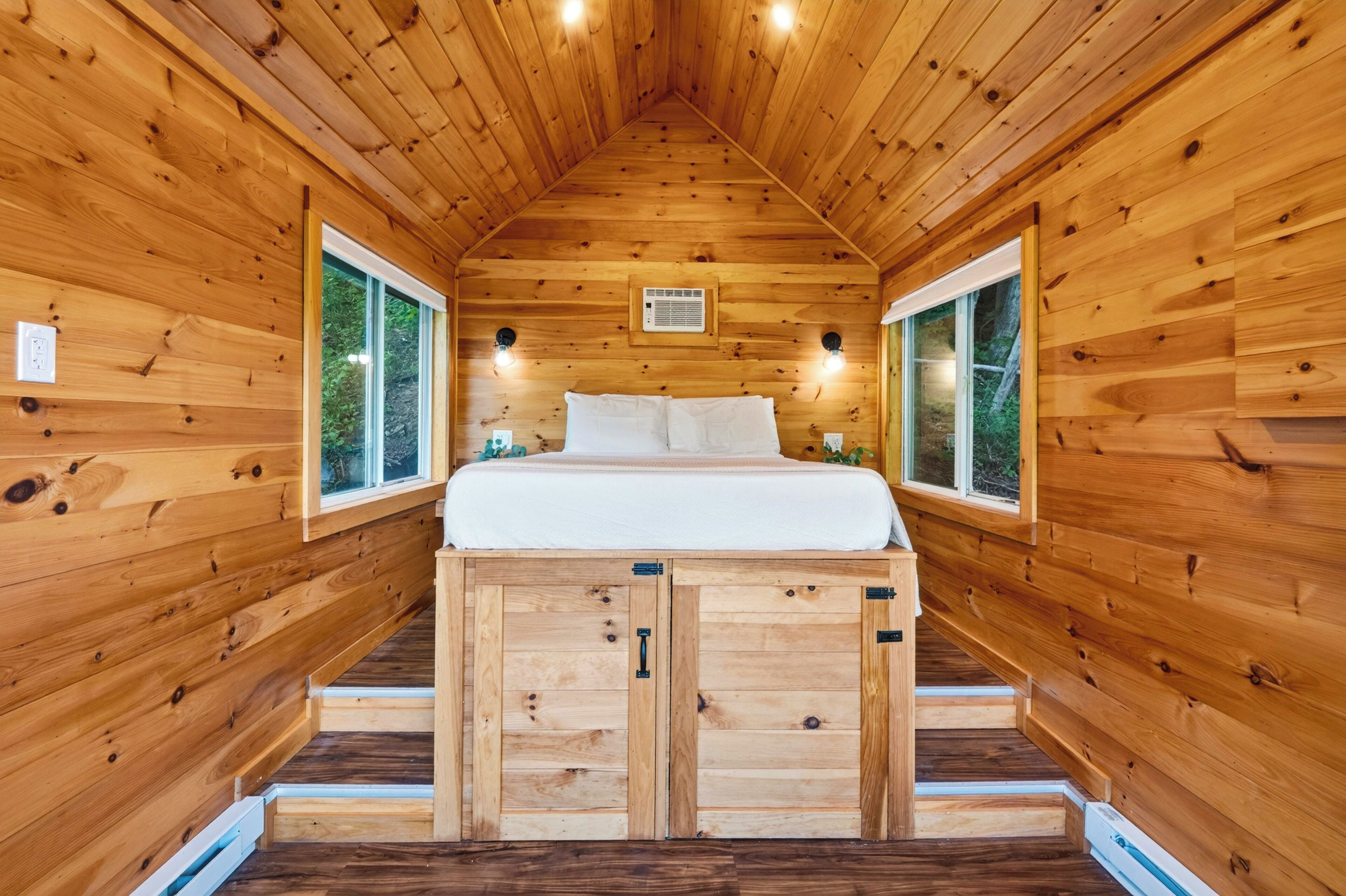 Una cabaña de madera con cama altillo, dos ventanas y un equipo de aire acondicionado pequeño.