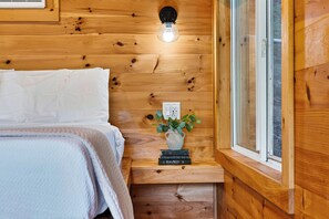 A wooden interior with a bed, a wall-mounted light, a small plant, and a window. - Firepit + Valley-View Deck | Creek + Valley Views + Slopes | Creekside Cabin (Lexington)