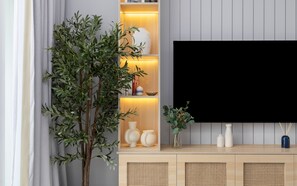 A living room with a large flat-screen TV mounted on a wall, a wooden cabinet with decorative items, and a potted plant. - Elegant Studio Retreat in Damac Hills (Dubai)