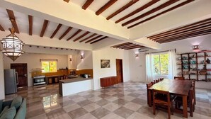 A spacious living area with a large wooden dining table, a kitchen in the background, and a checkered floor.