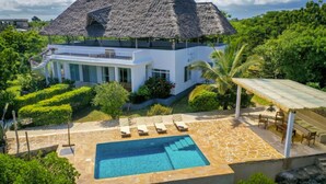 A modern villa with a pool, surrounded by lush greenery and a thatched-roof structure.