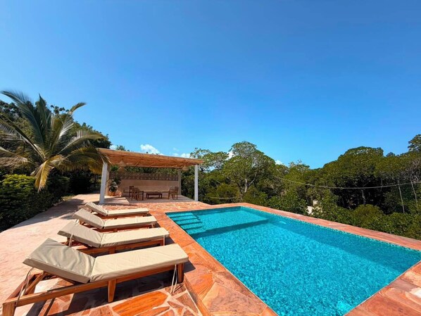 Piscina con sillones reclinables, una pérgola y vista a árboles.