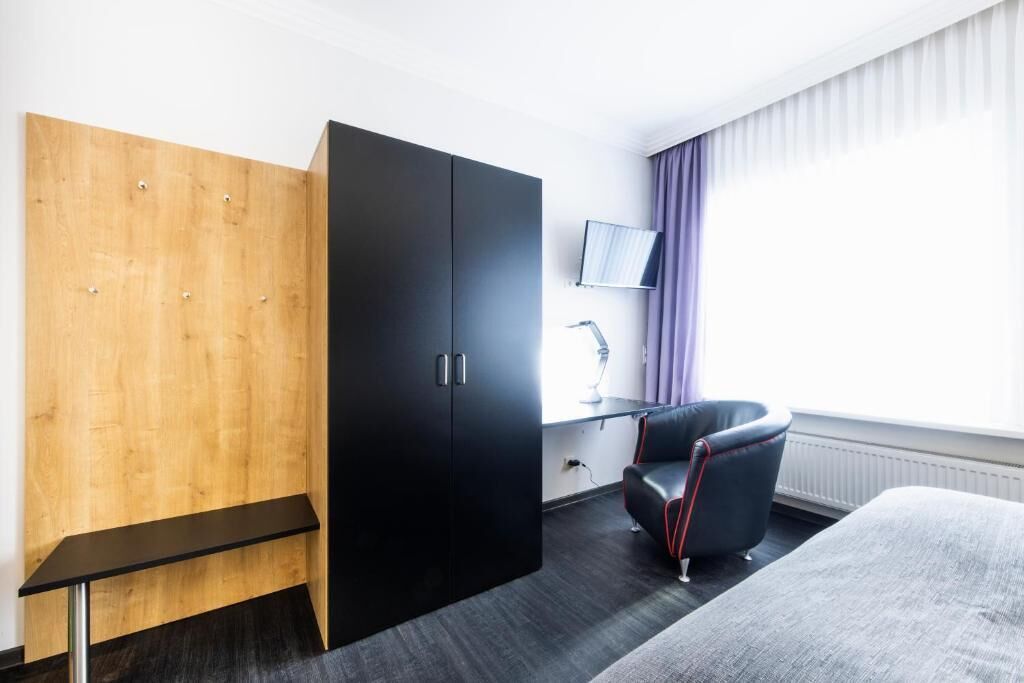Ein Hotelzimmer mit einem Holfter, einem schwarzen Schrank, einem Schreibtisch mit Lampe, einem schwarzen Ledersessel und einem Bett.