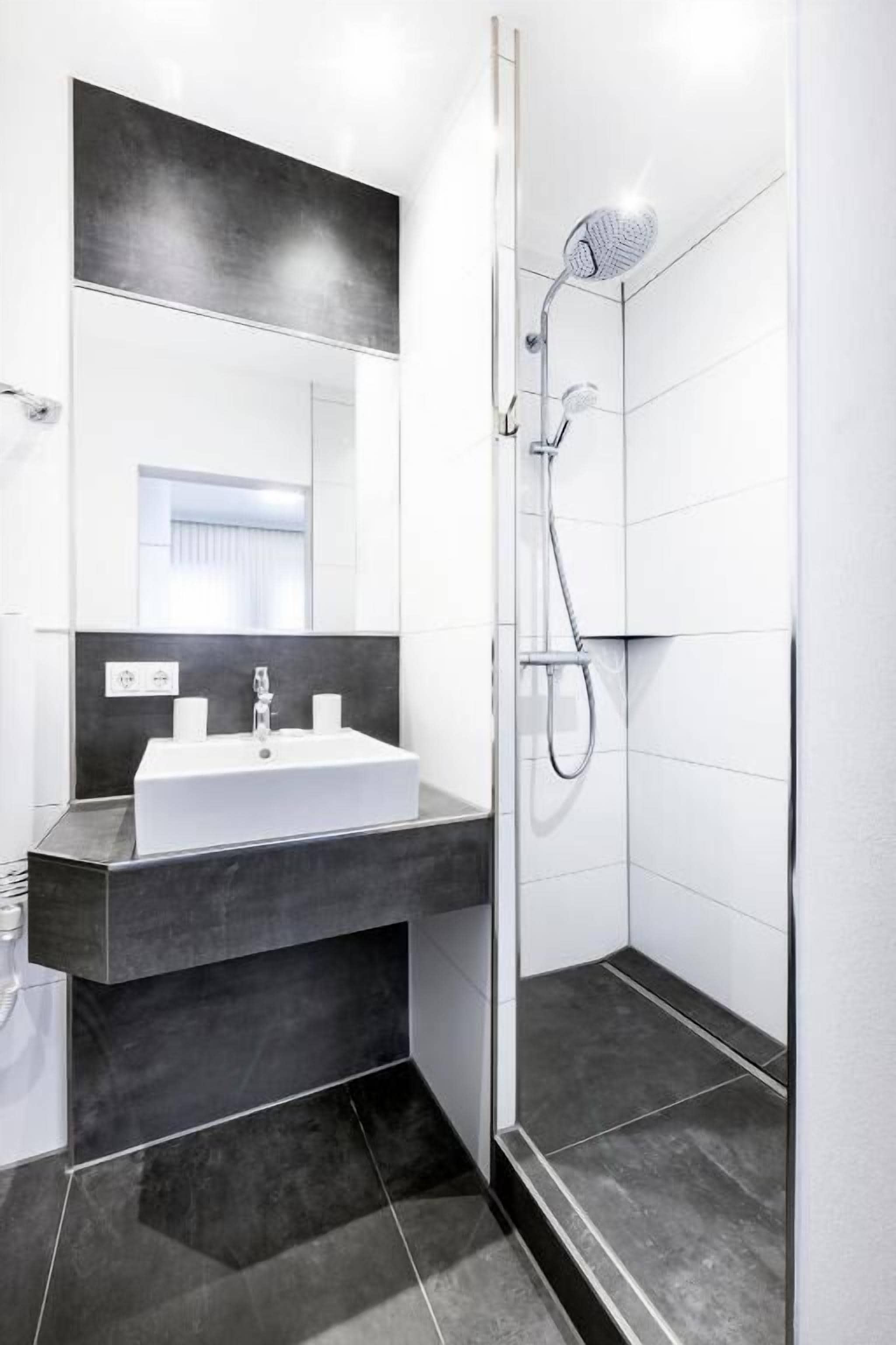 Un baño moderno con un lavamanos blanco, baldosas oscuras y una cabina de ducha de vidrio.