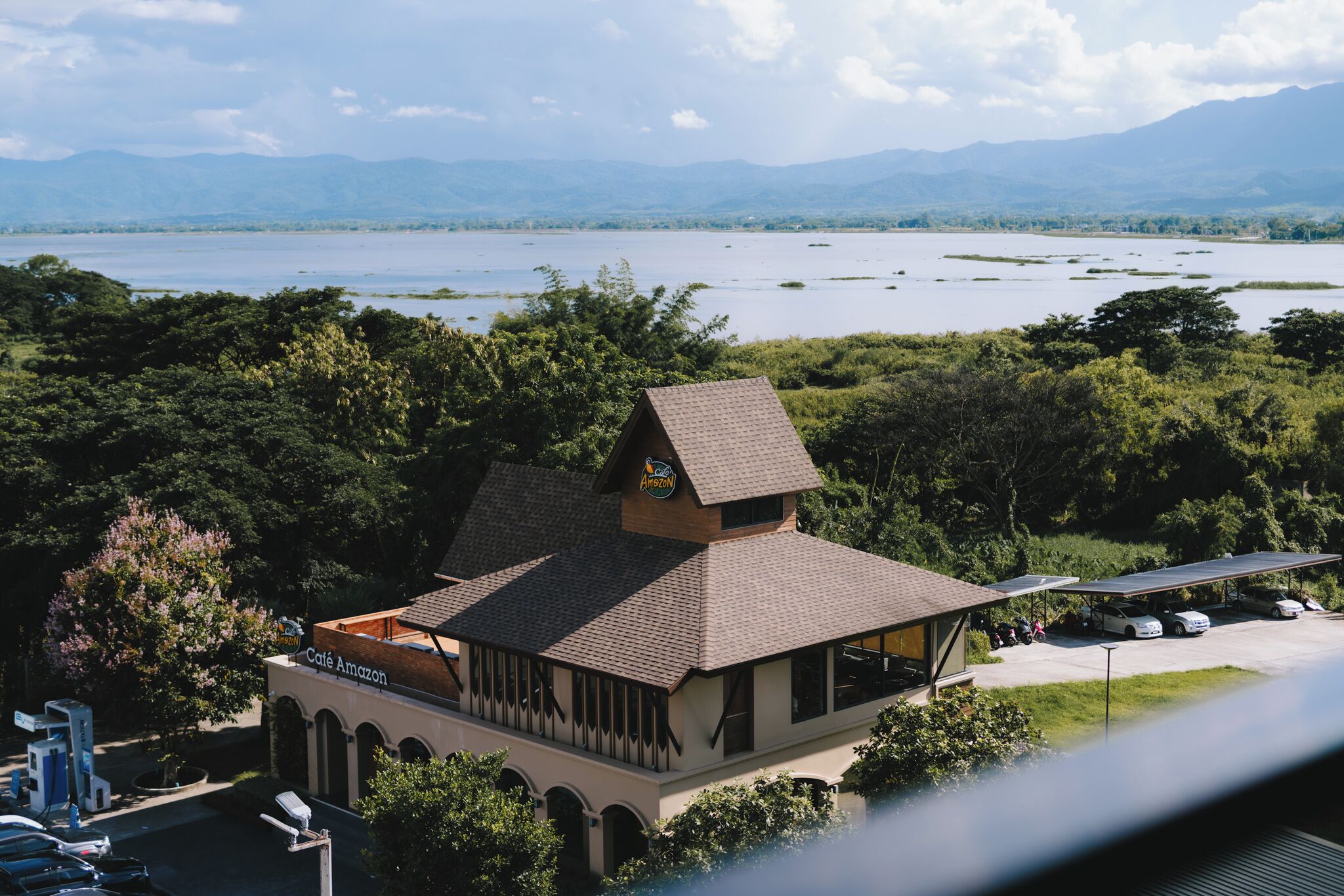 A building with a sloped roof and a café sign, surrounded by lush greenery and a lake.