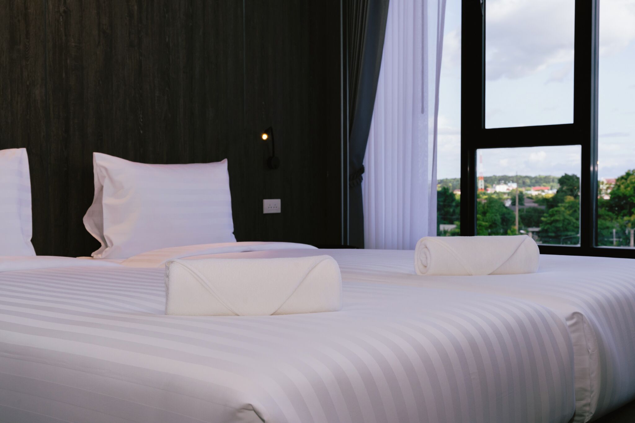 A double bed with white linens and pillows, a nightstand with a lamp, and a large window with a view of greenery and buildings.